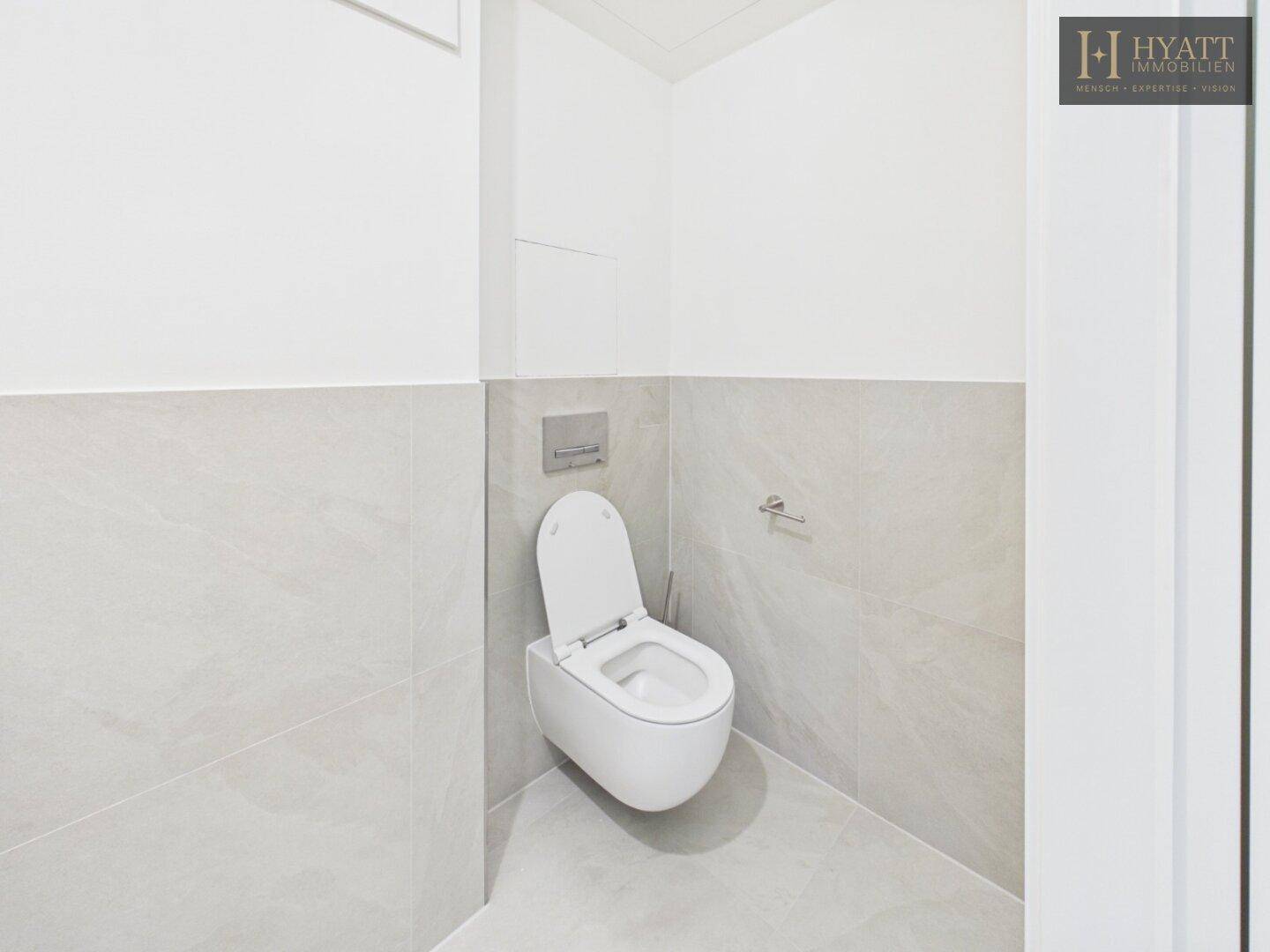 Modernes WC mit wandhängender Toilette und hellgrauen Fliesen an Wänden und Boden.
