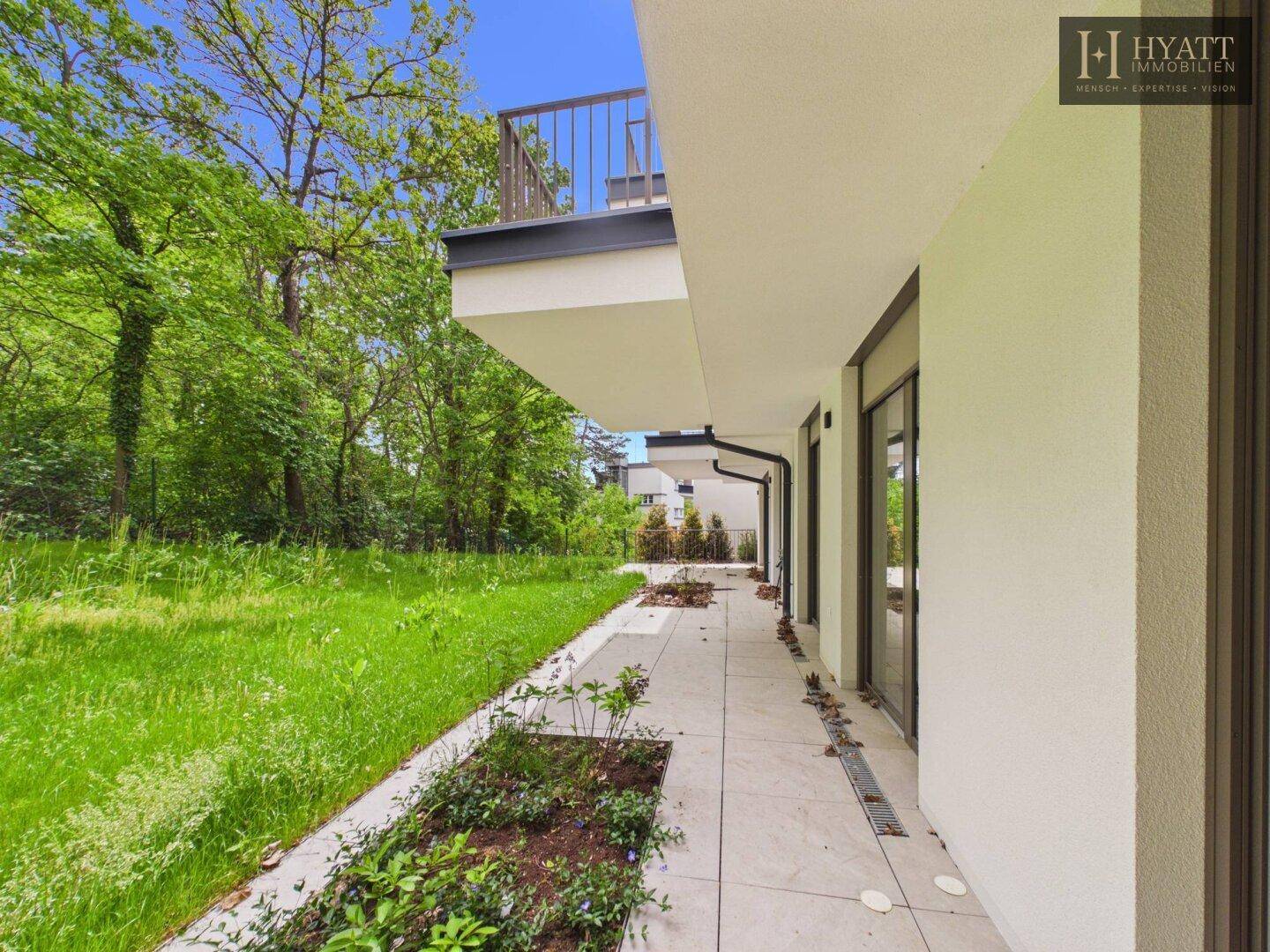 Lange, gepflasterte Terrasse entlang des Gebäudes mit Grünflächen und Blick ins Freie.