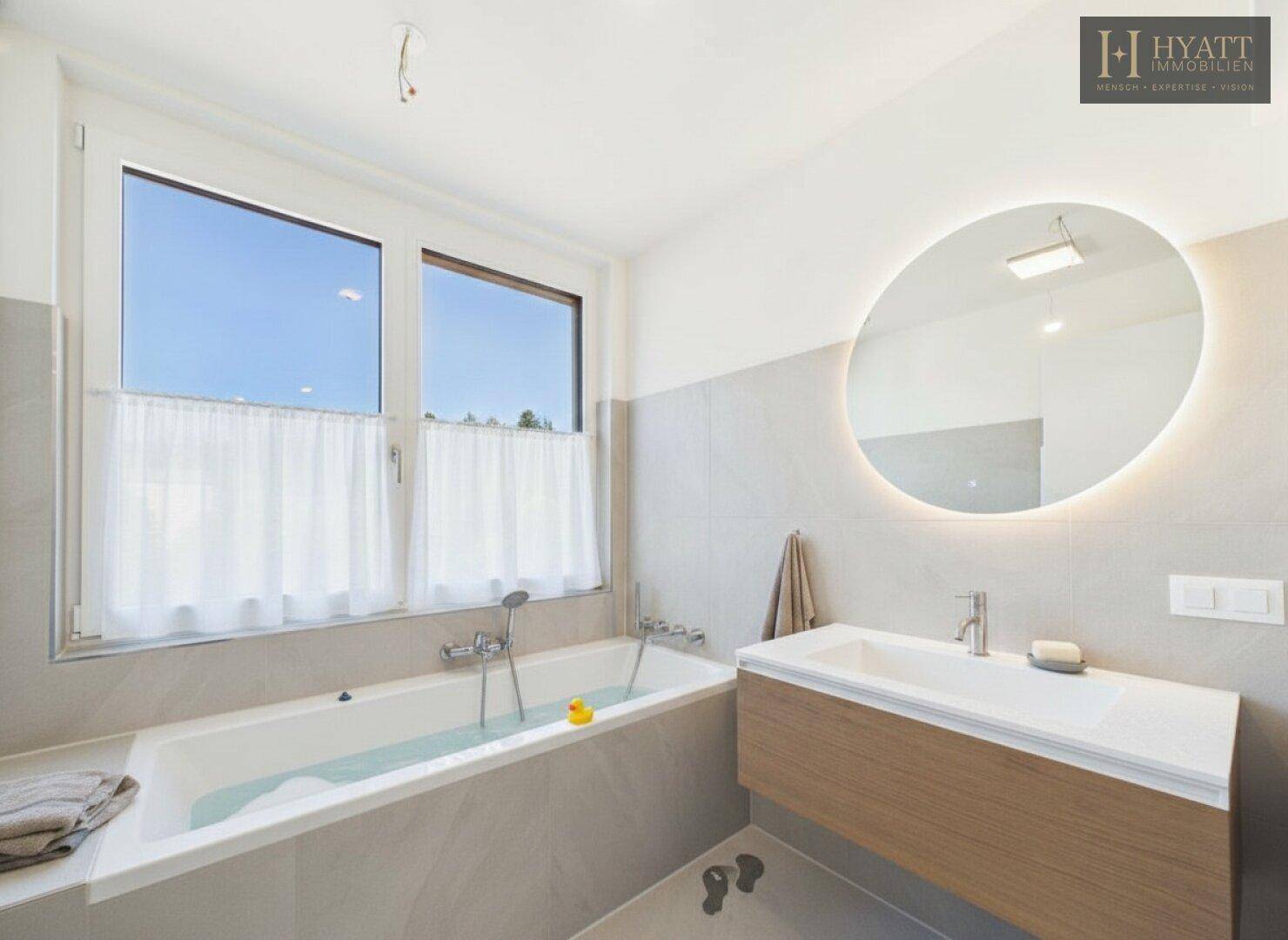 Modernes Badezimmer mit Badewanne, großem Fenster und beleuchtetem Spiegel.