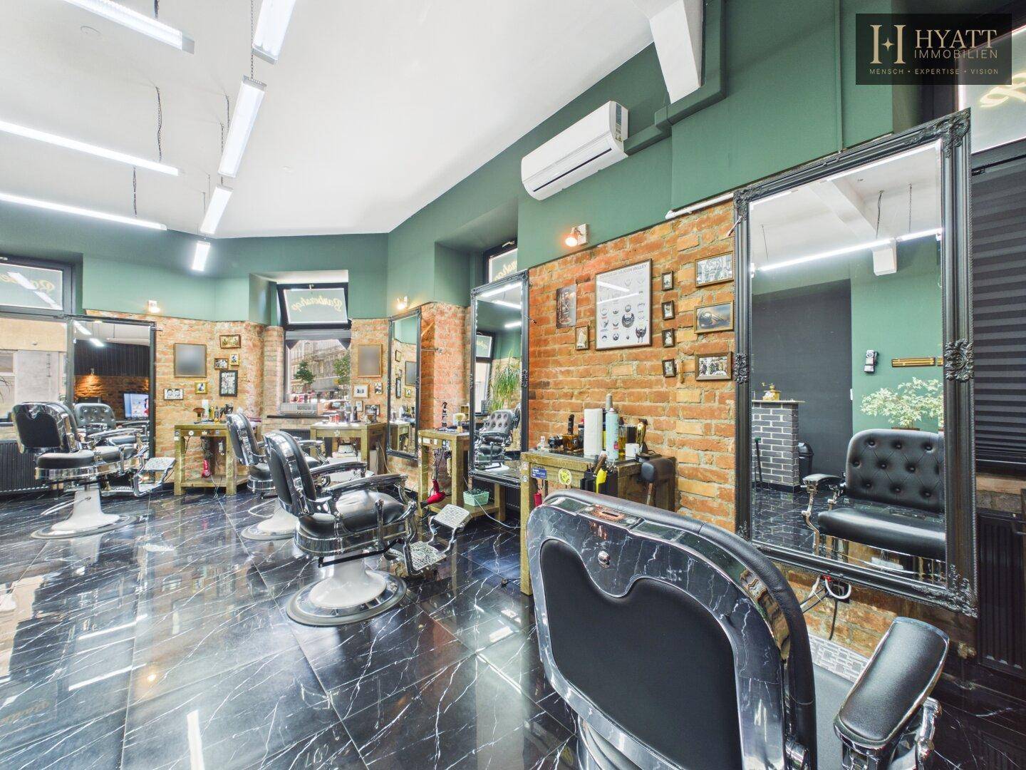 Blick in den stilvollen Friseursalon mit schwarzen Stühlen, großen Spiegeln und grünen Wänden.