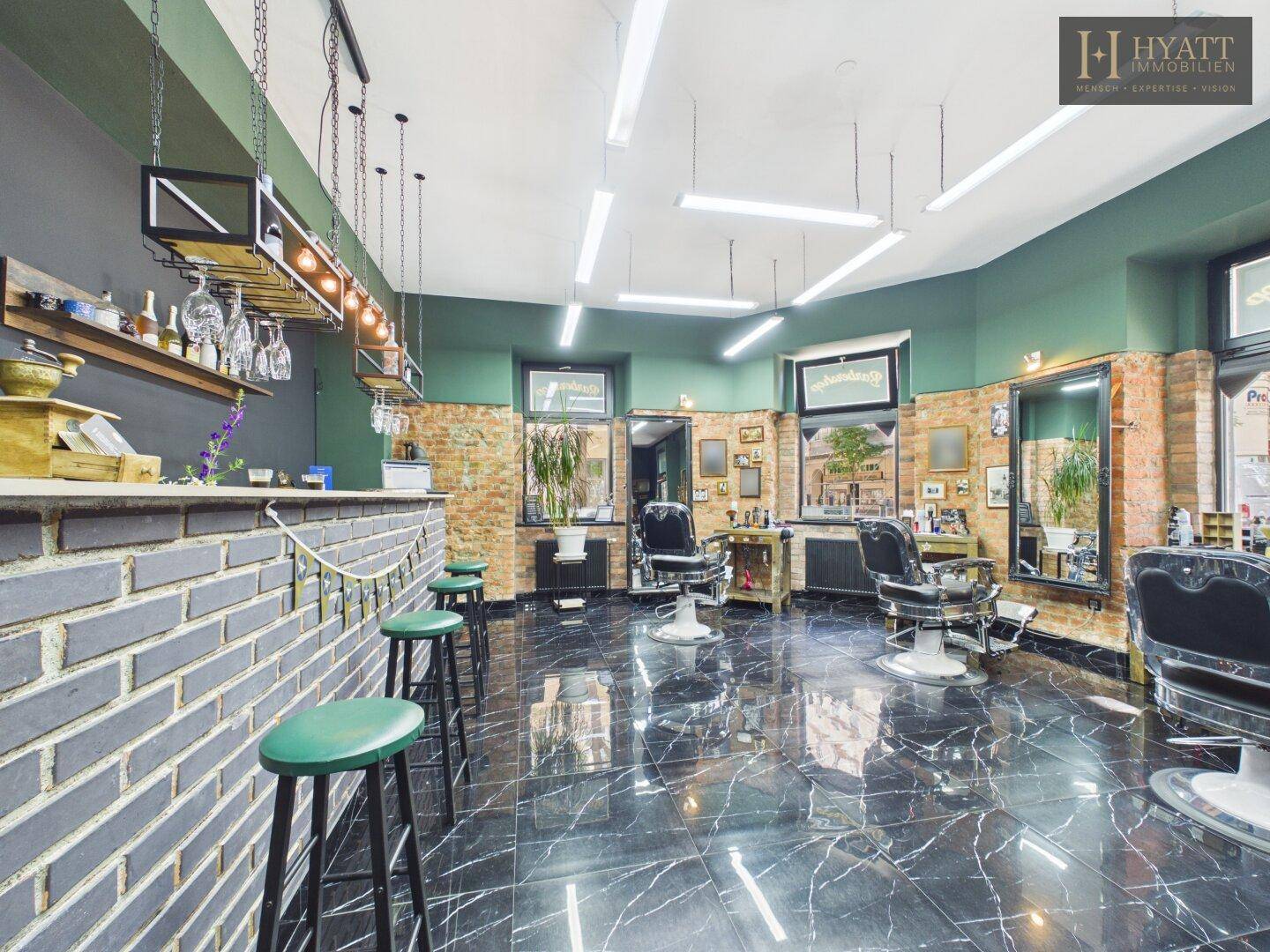 Barbereich im Friseursalon mit Ziegeltheke, grünen Barhockern und Hängeregal für Gläser.