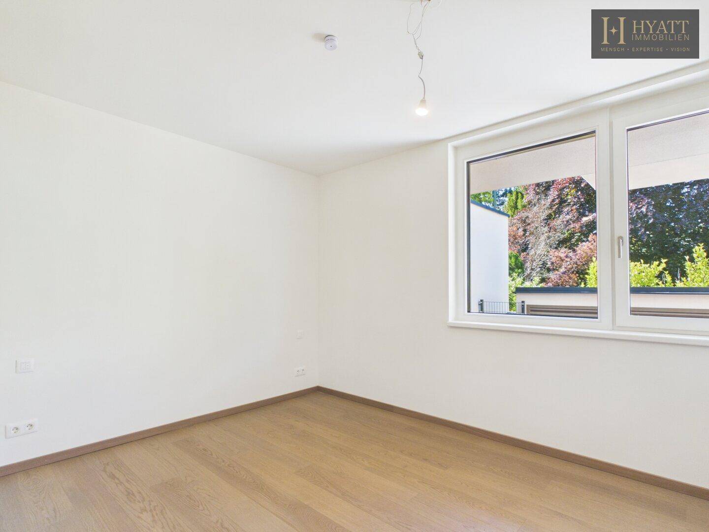 Helles Schlafzimmer mit Holzboden und großem Fenster mit Blick ins Grüne.