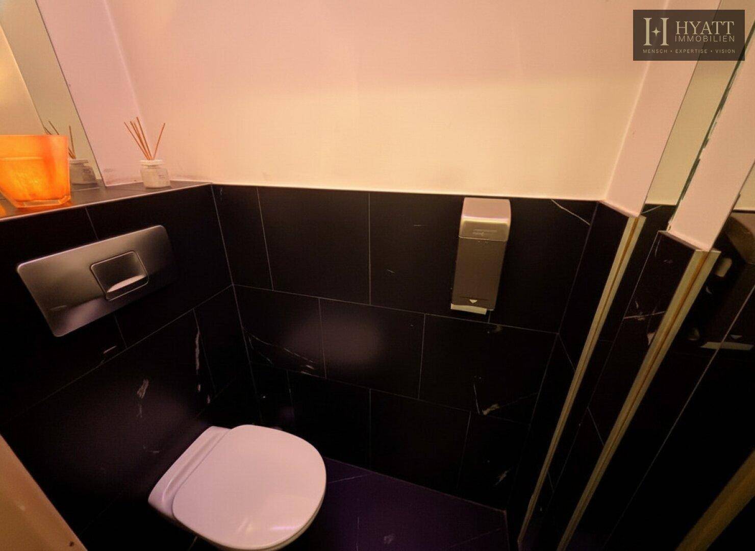 Saubere Toilettenkabine mit wandhängendem WC und dunklen, glänzenden Fliesen.