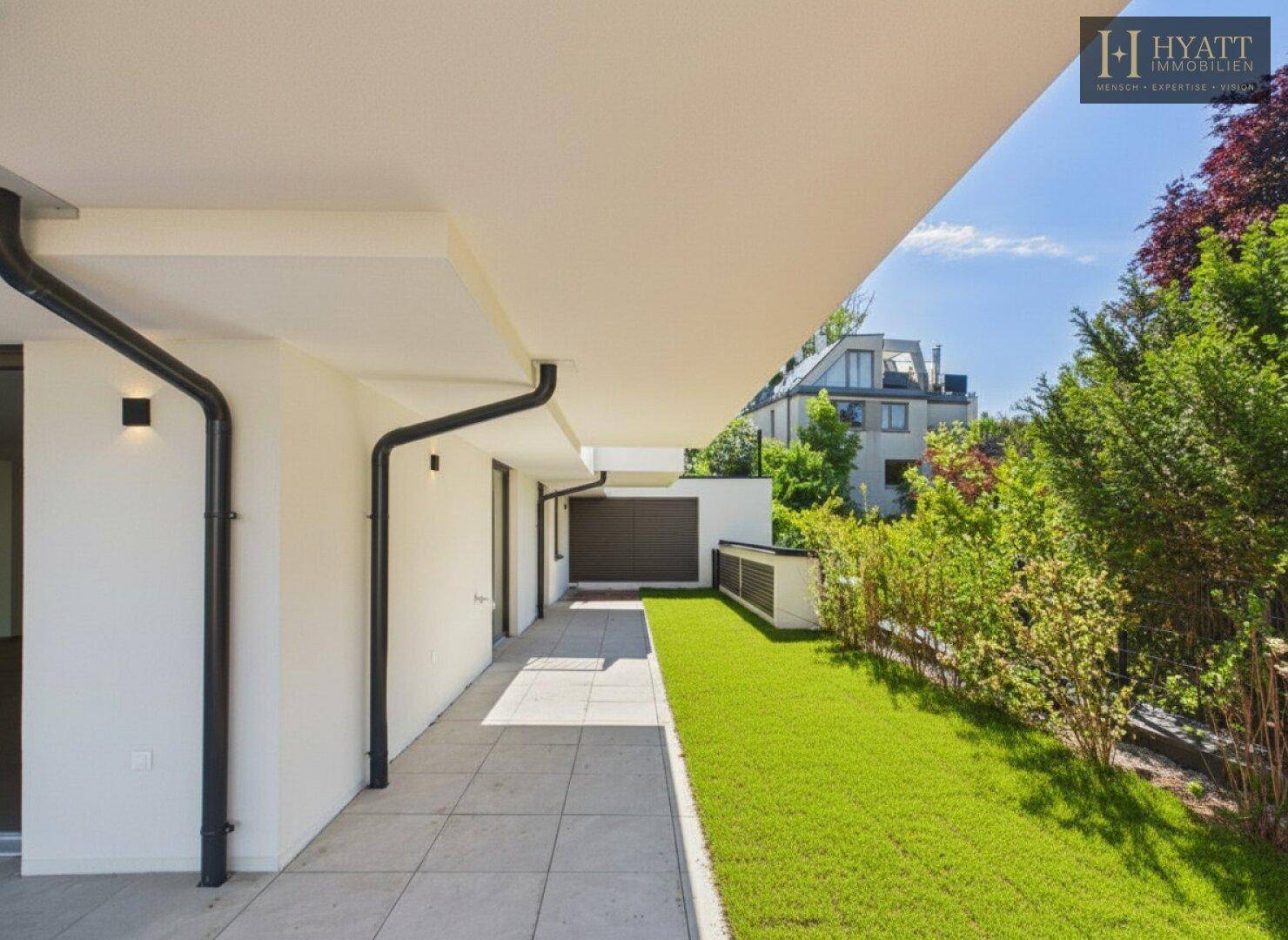 Lange, gepflasterte Terrasse entlang der Gebäudeseite mit schmalem Rasenstreifen und viel Grün.