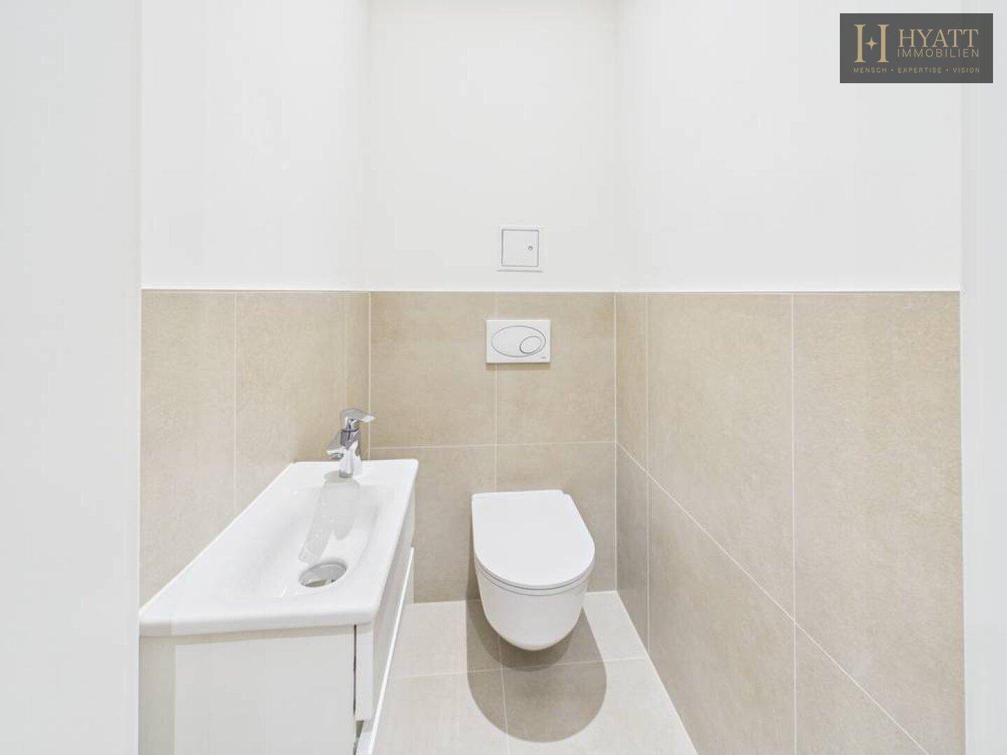 Separates WC mit wandhängender Toilette, kleinem Waschbecken und beigefarbenen Wand- und Bodenfliesen.