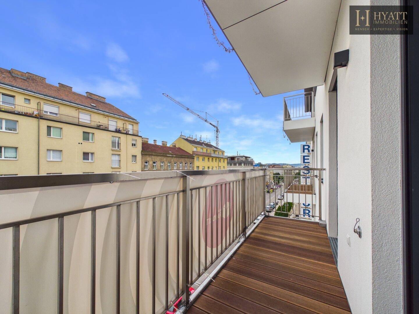 Geräumiger Balkon mit Holzboden und Blick auf die Stadt und umliegende Gebäude mit Kränen.