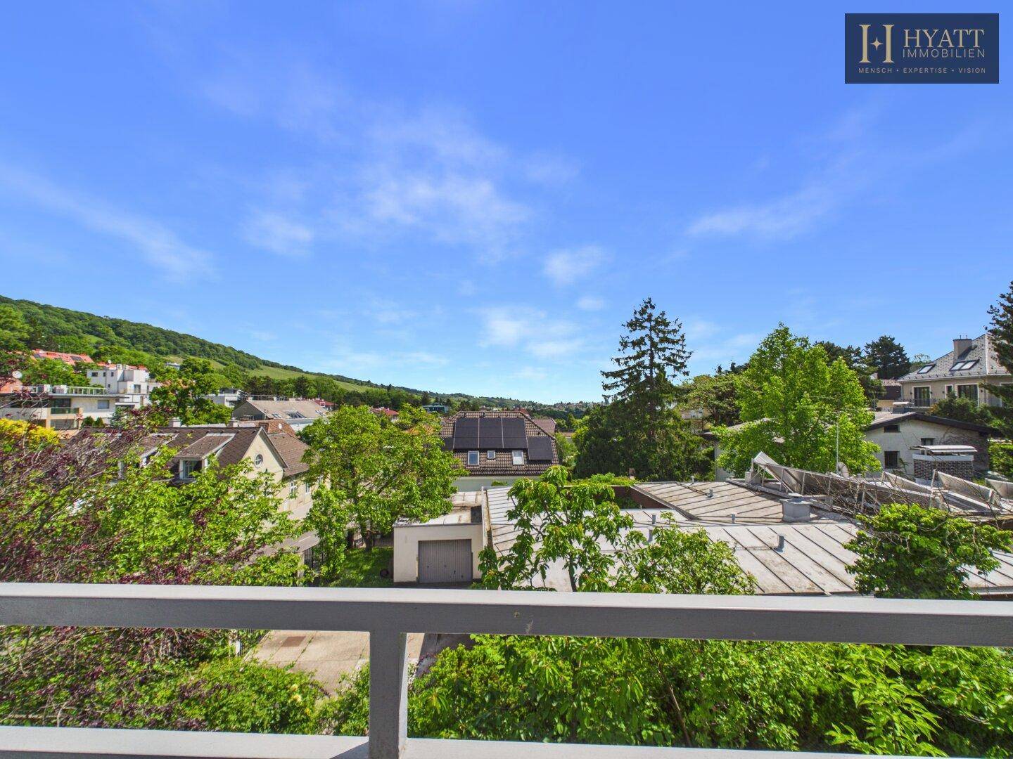 Weitblick von einem Balkon über eine grüne Wohngegend mit bewaldeten Hügeln im Hintergrund.