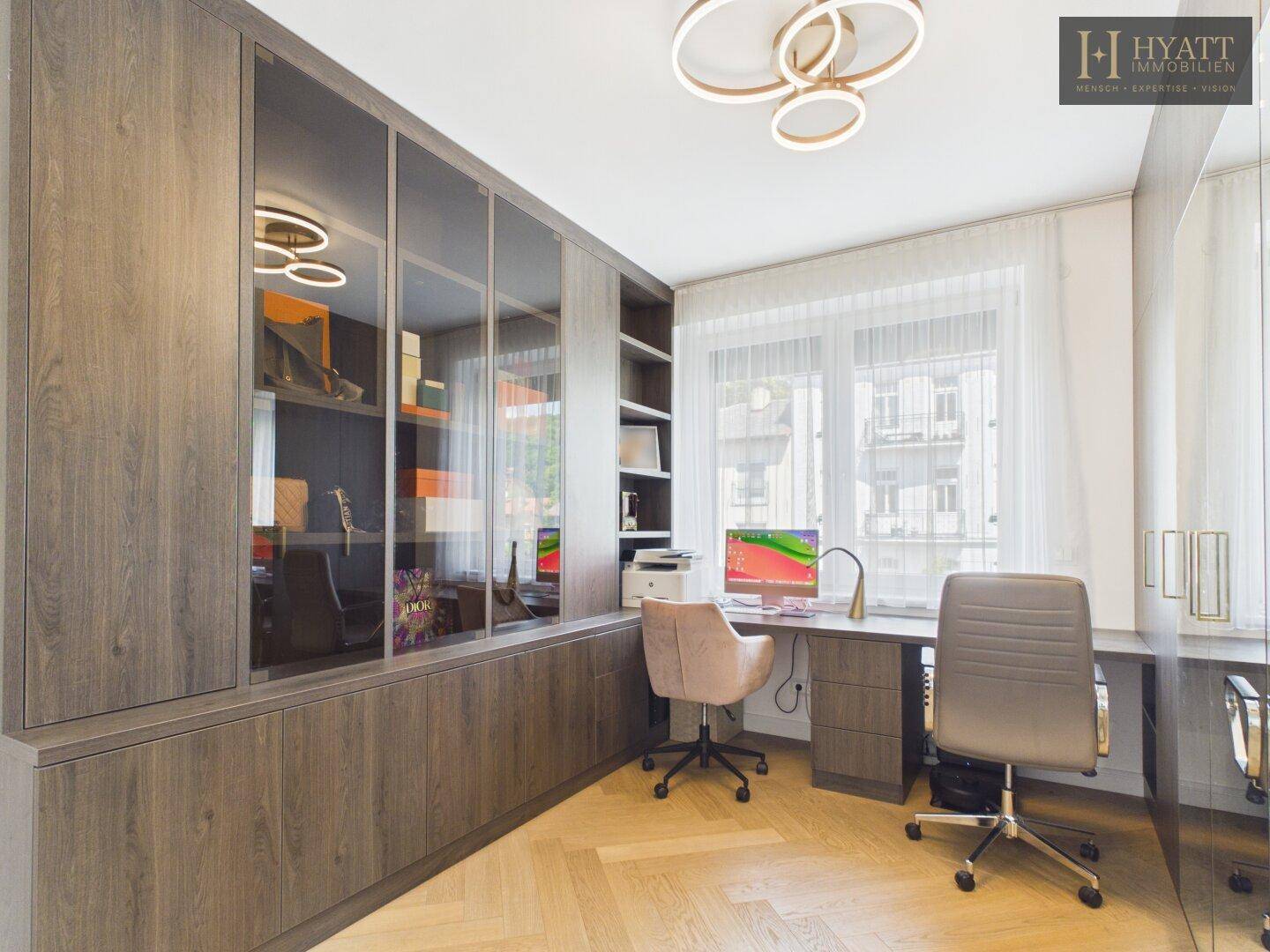 Helles Büro mit Einbauschränken, Holzparkett und zwei Arbeitsplätzen am Fenster.