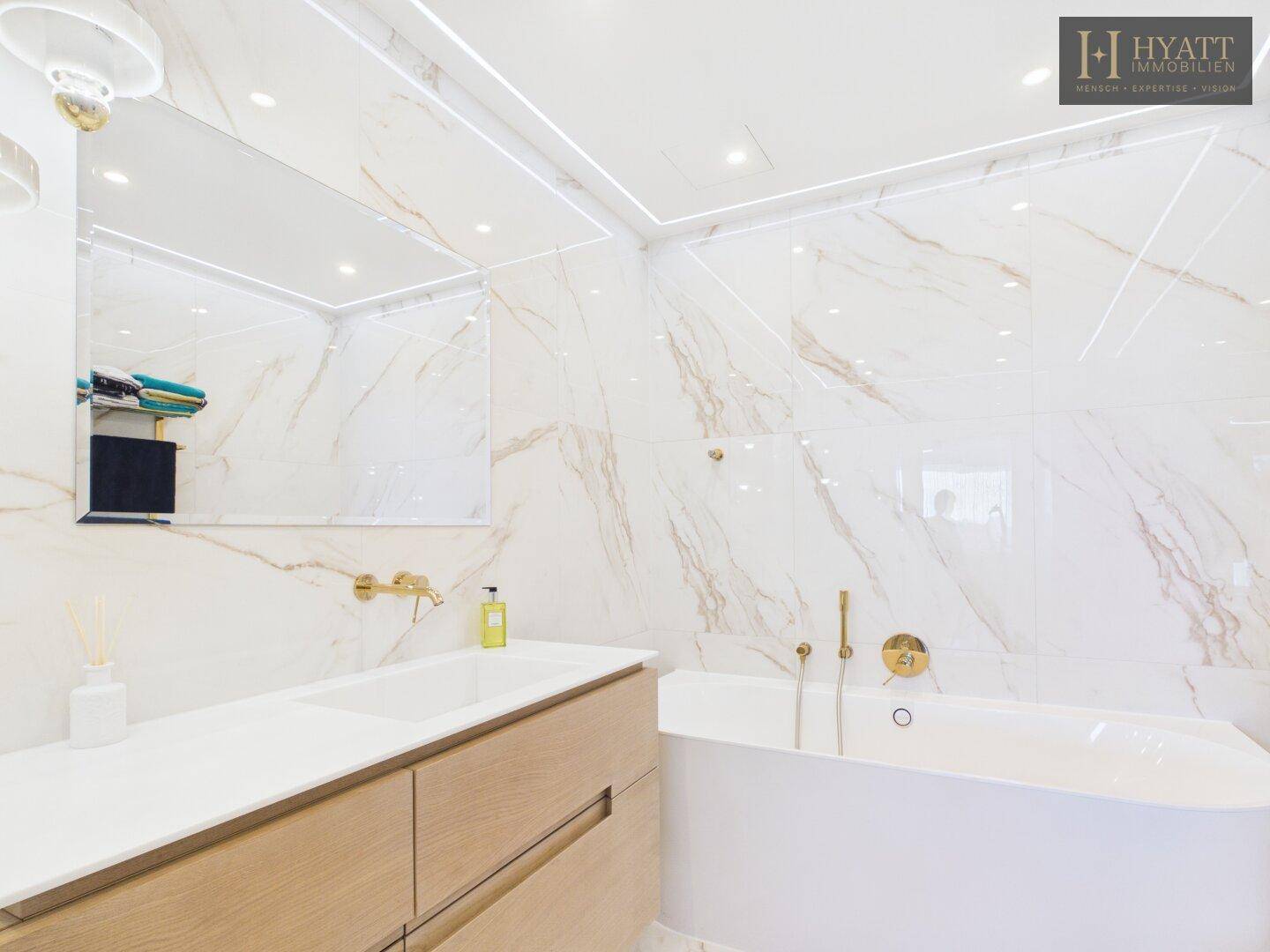 Luxuriöses Badezimmer mit Badewanne, Marmorfliesen und großem Spiegel mit integrierter Beleuchtung.