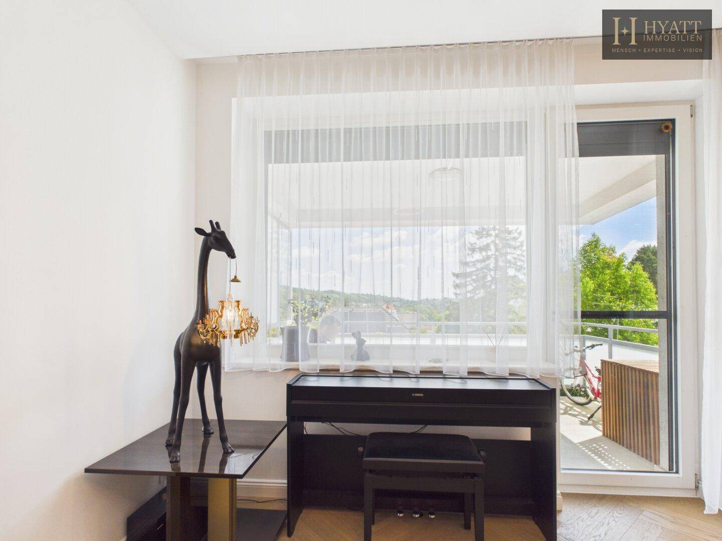 Wohnzimmer mit Klavier, Blick ins Grüne und Zugang zu einem sonnigen Balkon.