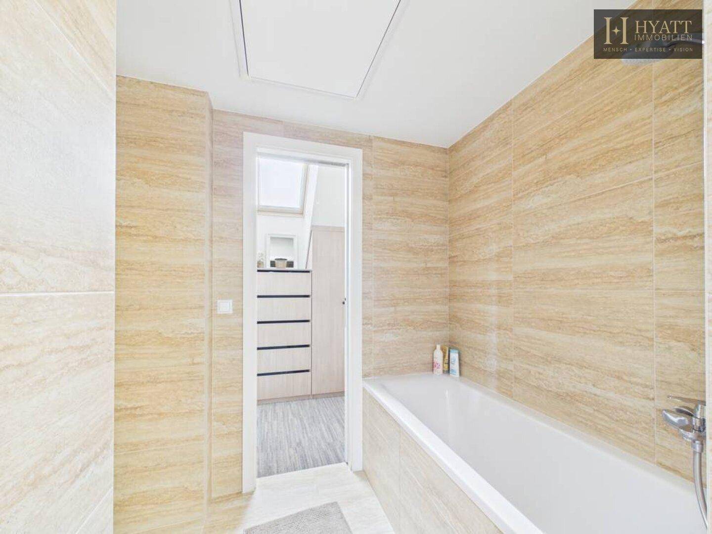 Helles Badezimmer mit Badewanne und beigefarbenen Fliesen, verbunden mit einem Ankleidebereich.