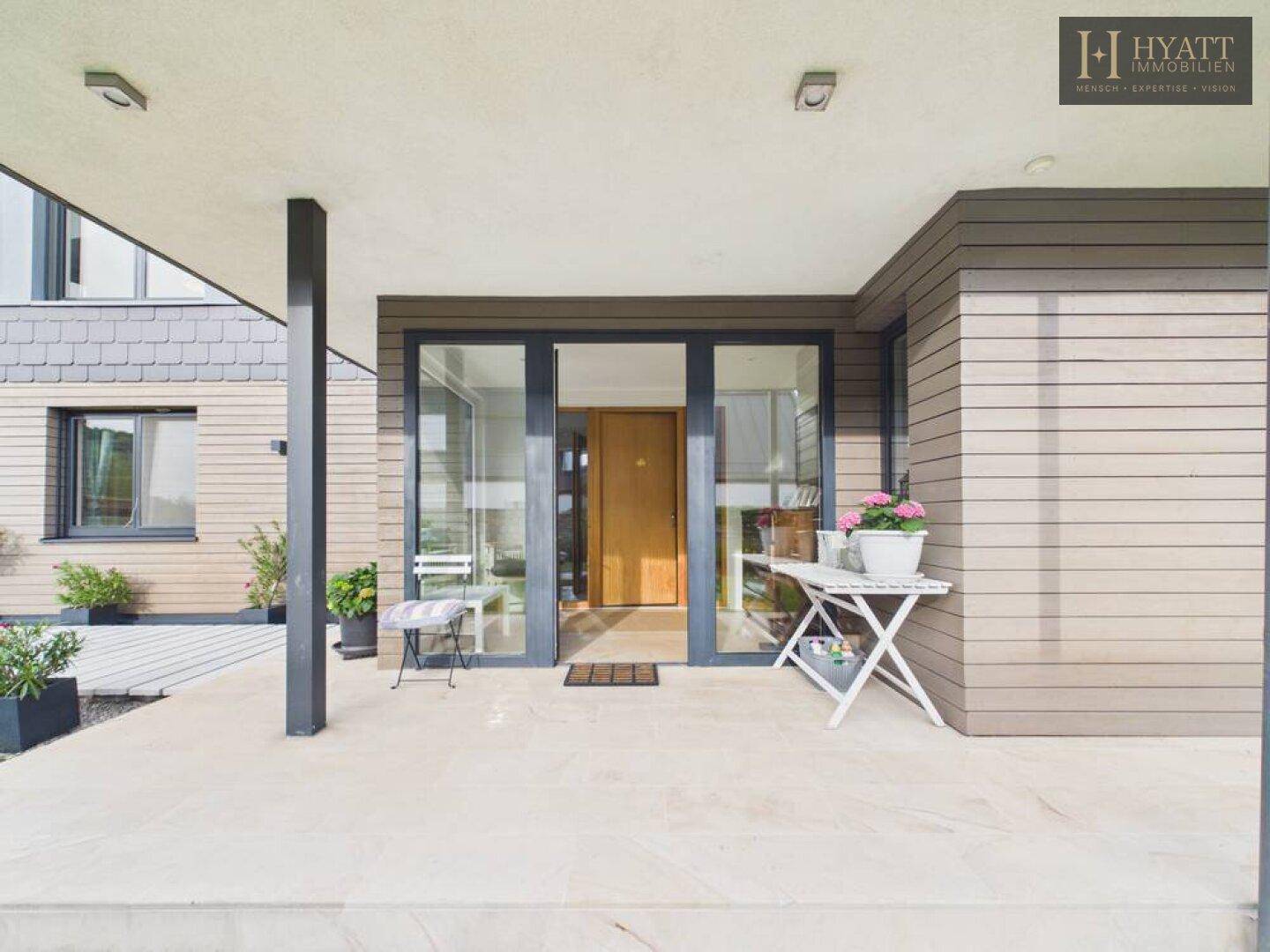 Überdachter Eingangsbereich des Hauses mit großen Glastüren und moderner Fassade.