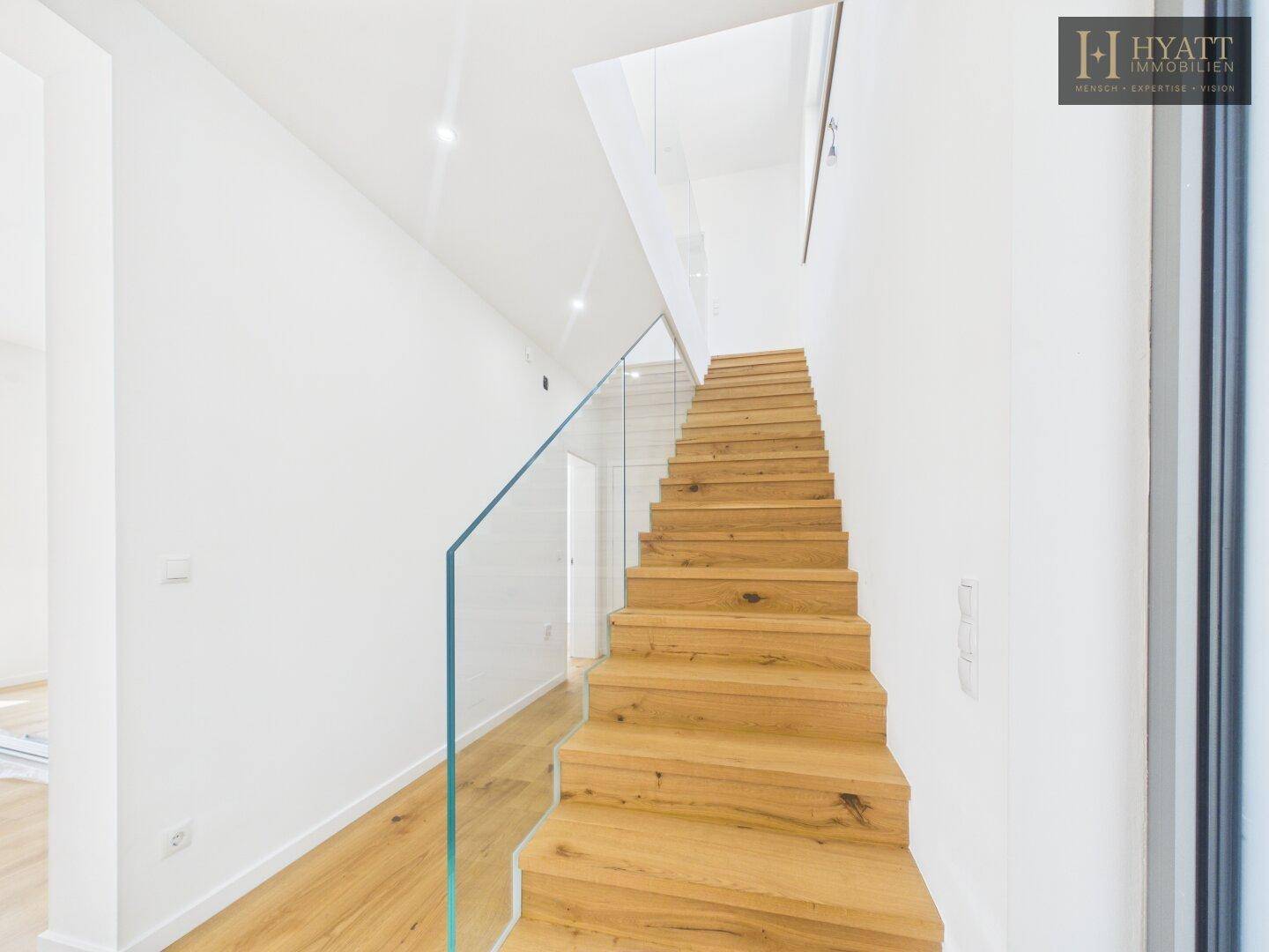 Moderne Holztreppe mit Glasgeländer in einem hellen, offenen Treppenhaus.