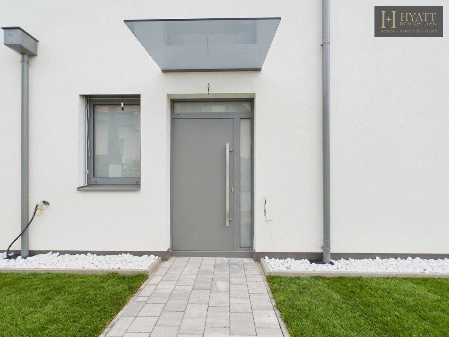 Nahaufnahme des modernen Hauseingangs mit grauer Tür, Glasvordach und weißem Kiesbeet.