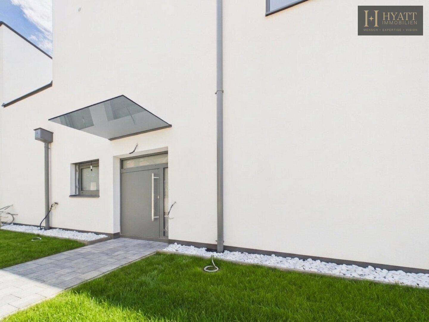 Moderner Hauseingang mit grauer Tür, Glasvordach und gepflegtem Vorgartenbereich.