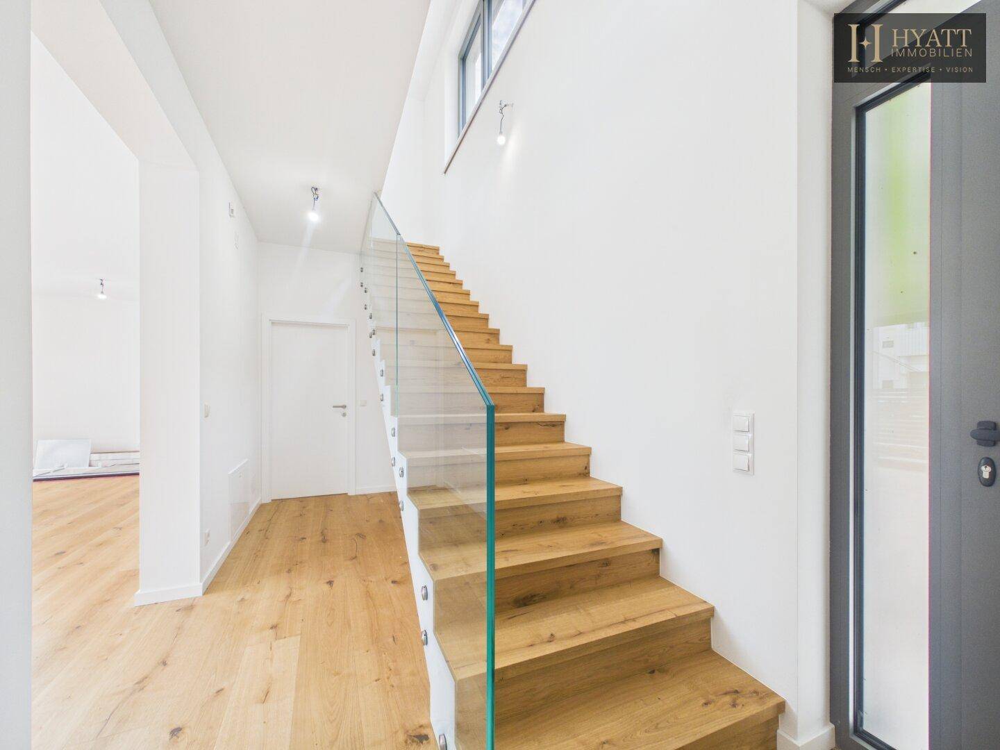 Heller Eingangsbereich mit moderner Holztreppe, Glasgeländer und viel natürlichem Licht.