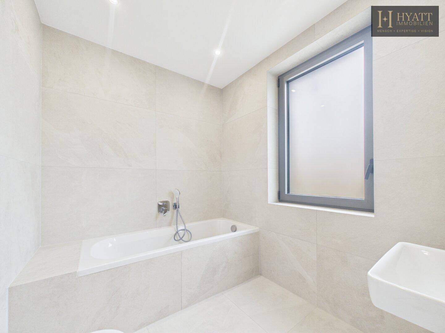 Modernes Badezimmer mit Badewanne, Waschbecken und Fenster, hell gefliest.