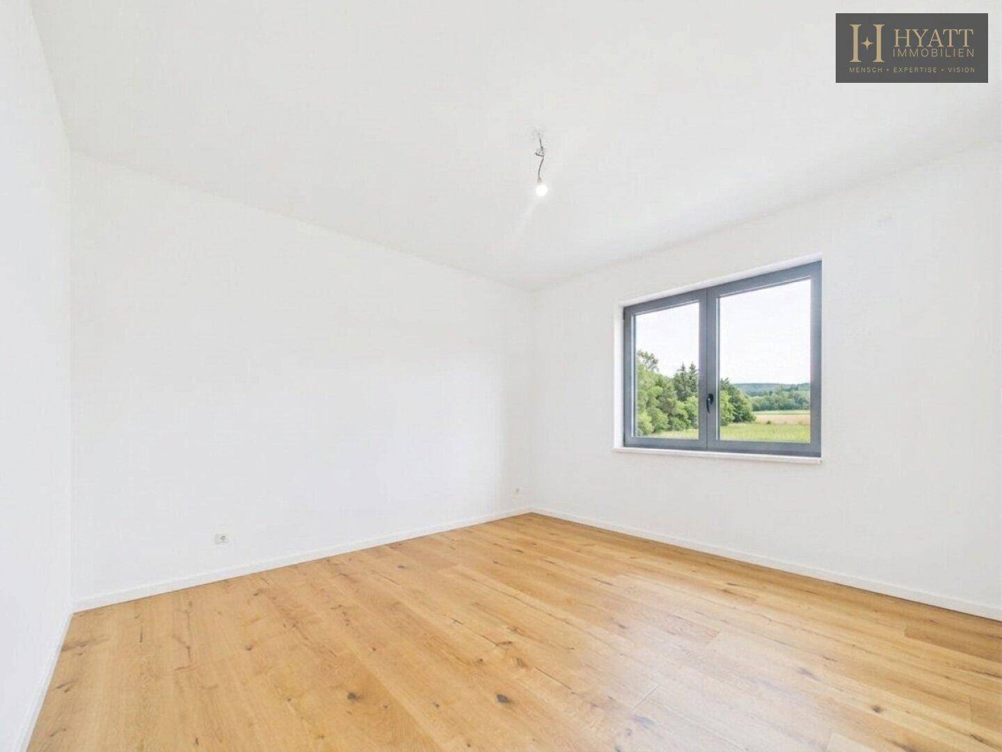 Leeres Schlafzimmer mit Holzboden und Fenster, das einen ruhigen Blick ins Grüne bietet.