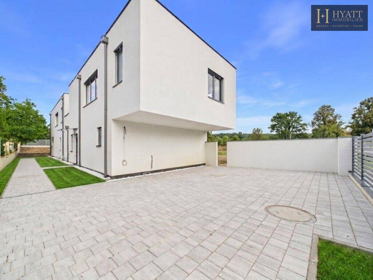 Rückseite des modernen Hauses mit gepflastertem Parkplatz und seitlichem Grünstreifen.