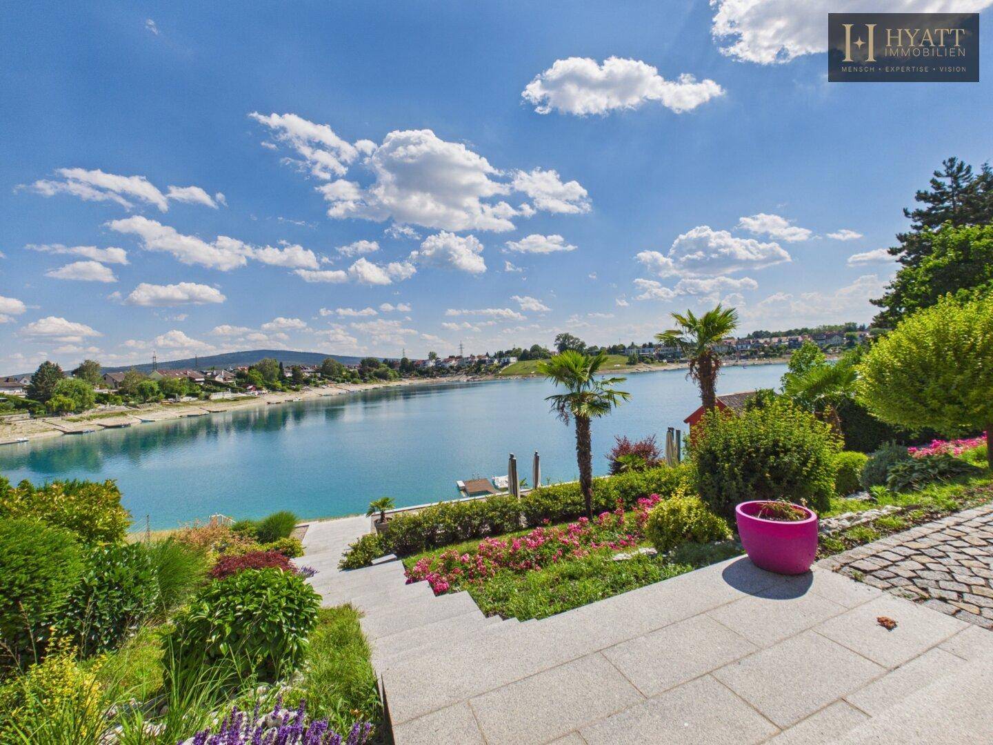 Gepflegter Garten mit Treppen, blühenden Pflanzen, Palmen und direktem Zugang zum See.