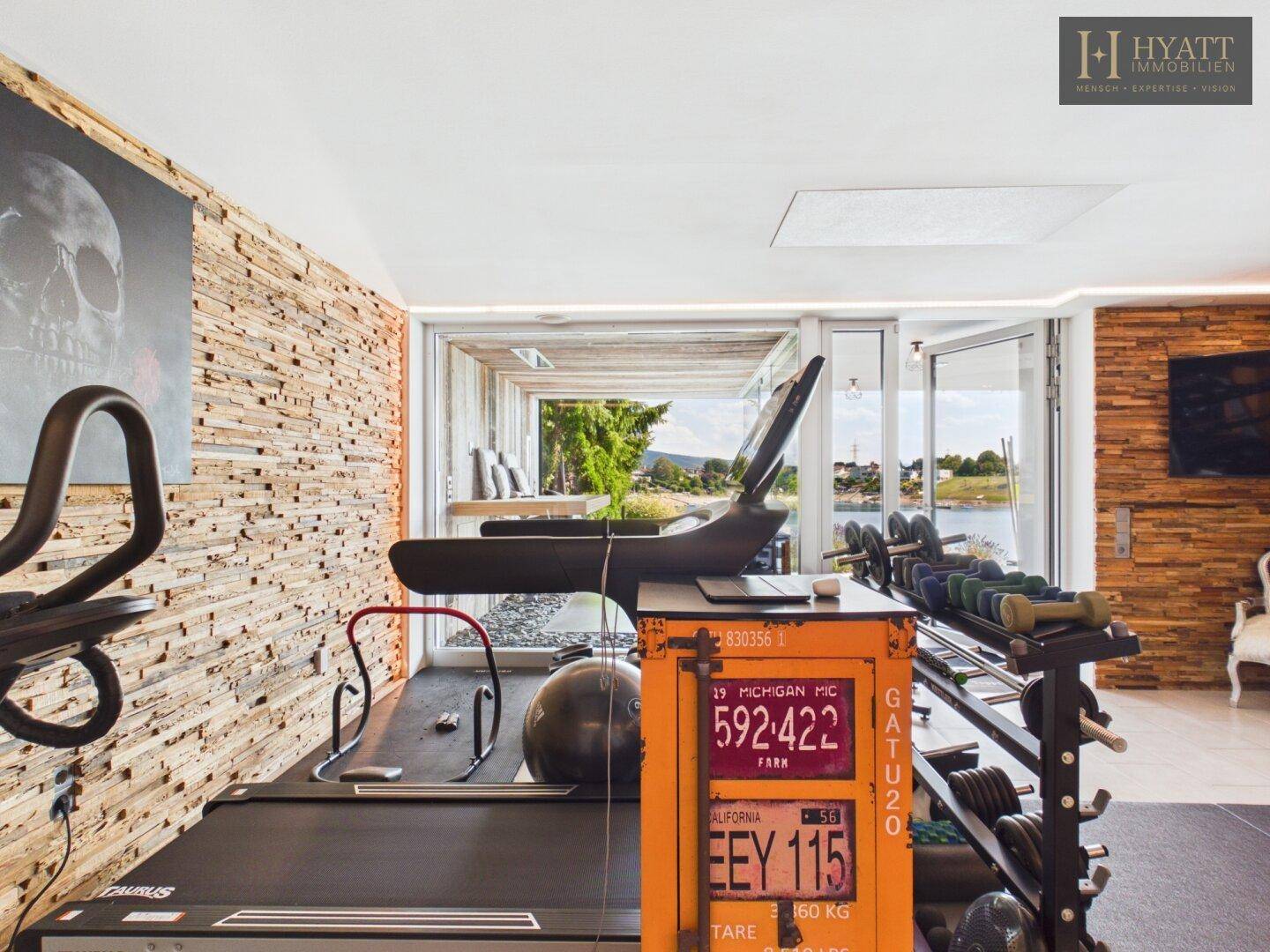 Moderner Fitnessraum mit Trainingsgeräten, Holzwänden und Panoramafenstern zum See.
