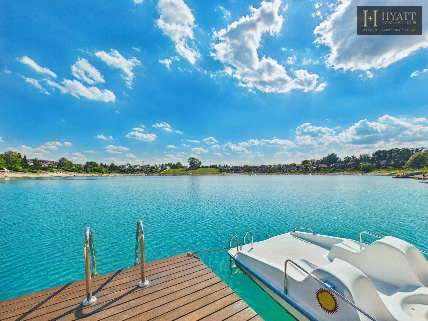 Holzsteg mit Tretboot am Ufer eines klaren, blauen Sees unter strahlendem Himmel.