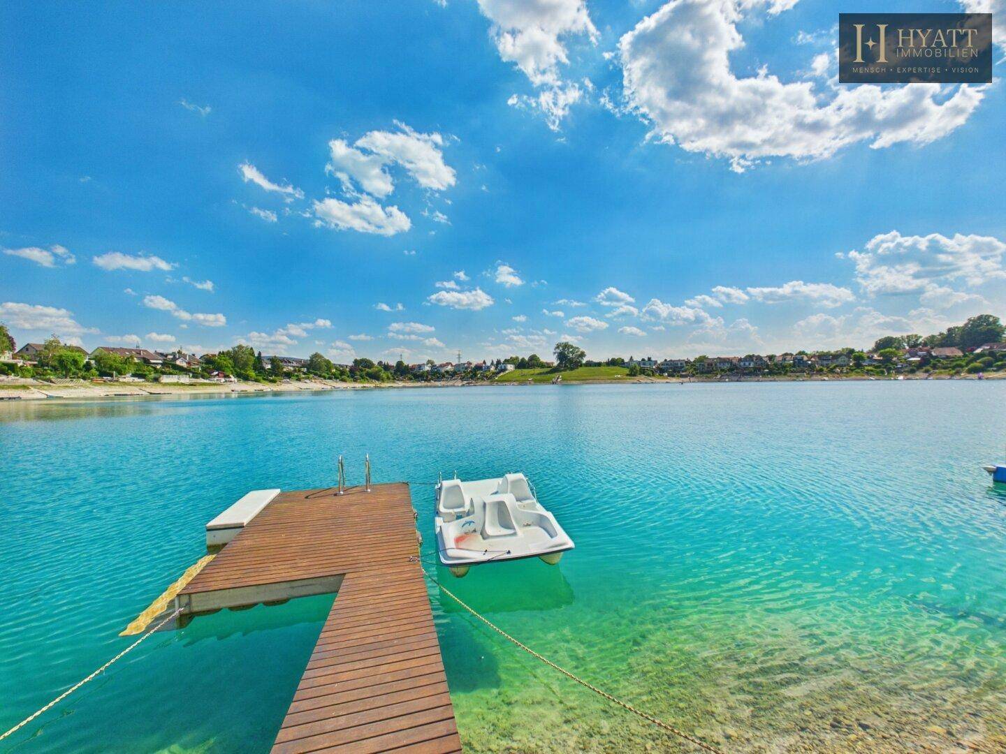 Langer Holzsteg mit Tretboot im türkisfarbenen See, umgeben von einer ruhigen Landschaft.