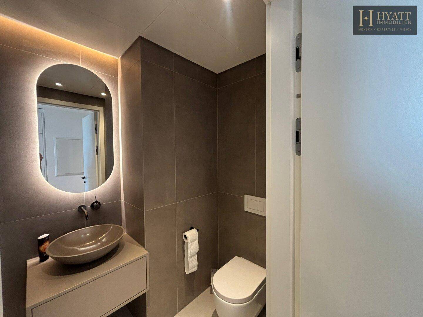 Modernes WC mit Wand-WC, Aufsatzwaschbecken und beleuchtetem Spiegel in grauen Fliesen.