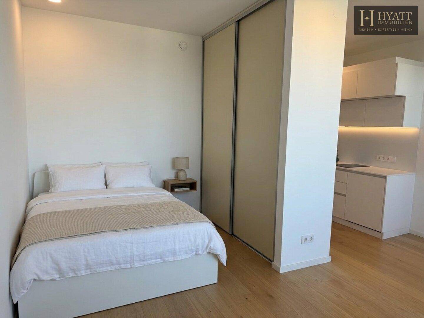 Schlafzimmer mit Bett, Nachttisch und integriertem Kleiderschrank, angrenzend an eine Küchenzeile.