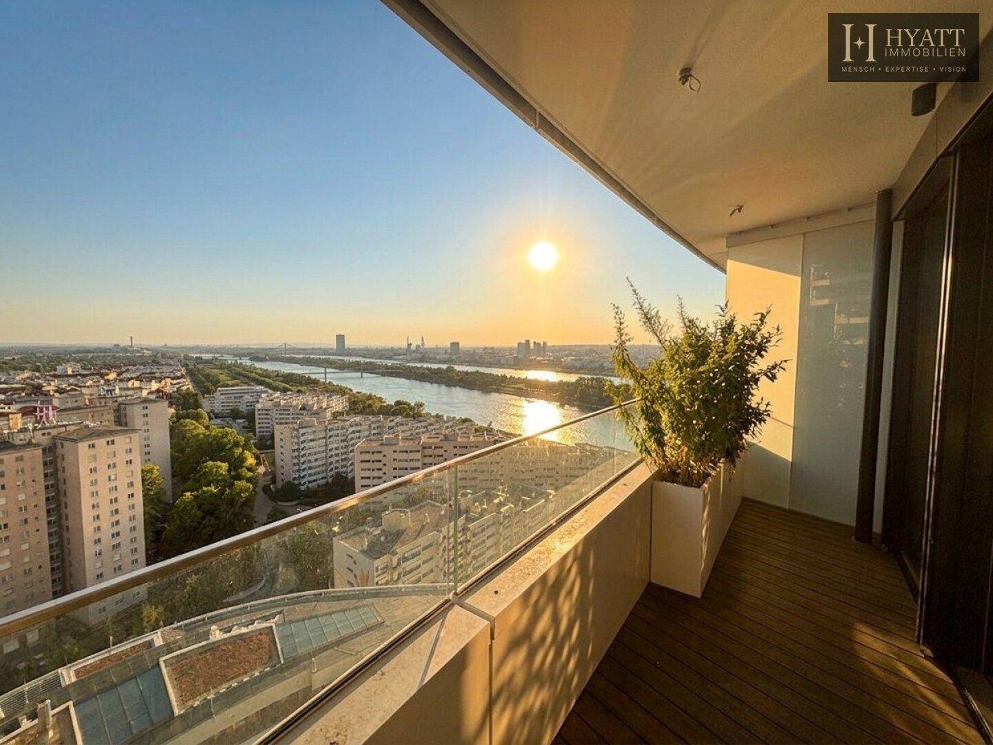 Balkon mit Glasgeländer und atemberaubendem Blick auf die Stadt und den Fluss bei Sonnenuntergang.