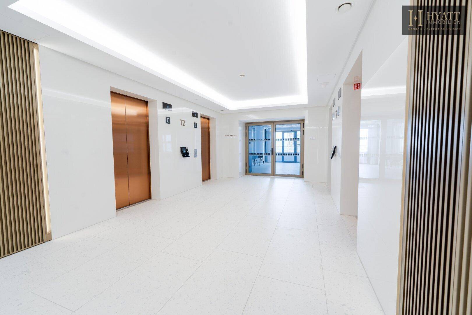 Moderner Flur mit Aufzügen, hellen Fliesenboden und indirekter Deckenbeleuchtung in einem Neubau.