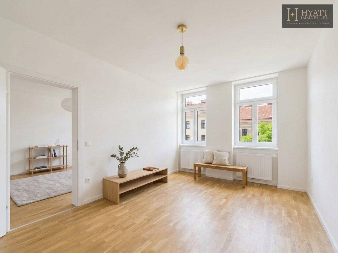 Lichtdurchfluteter Raum mit Parkettboden, großen Fenstern und offenem Durchgang zu einem angrenzenden Zimmer.