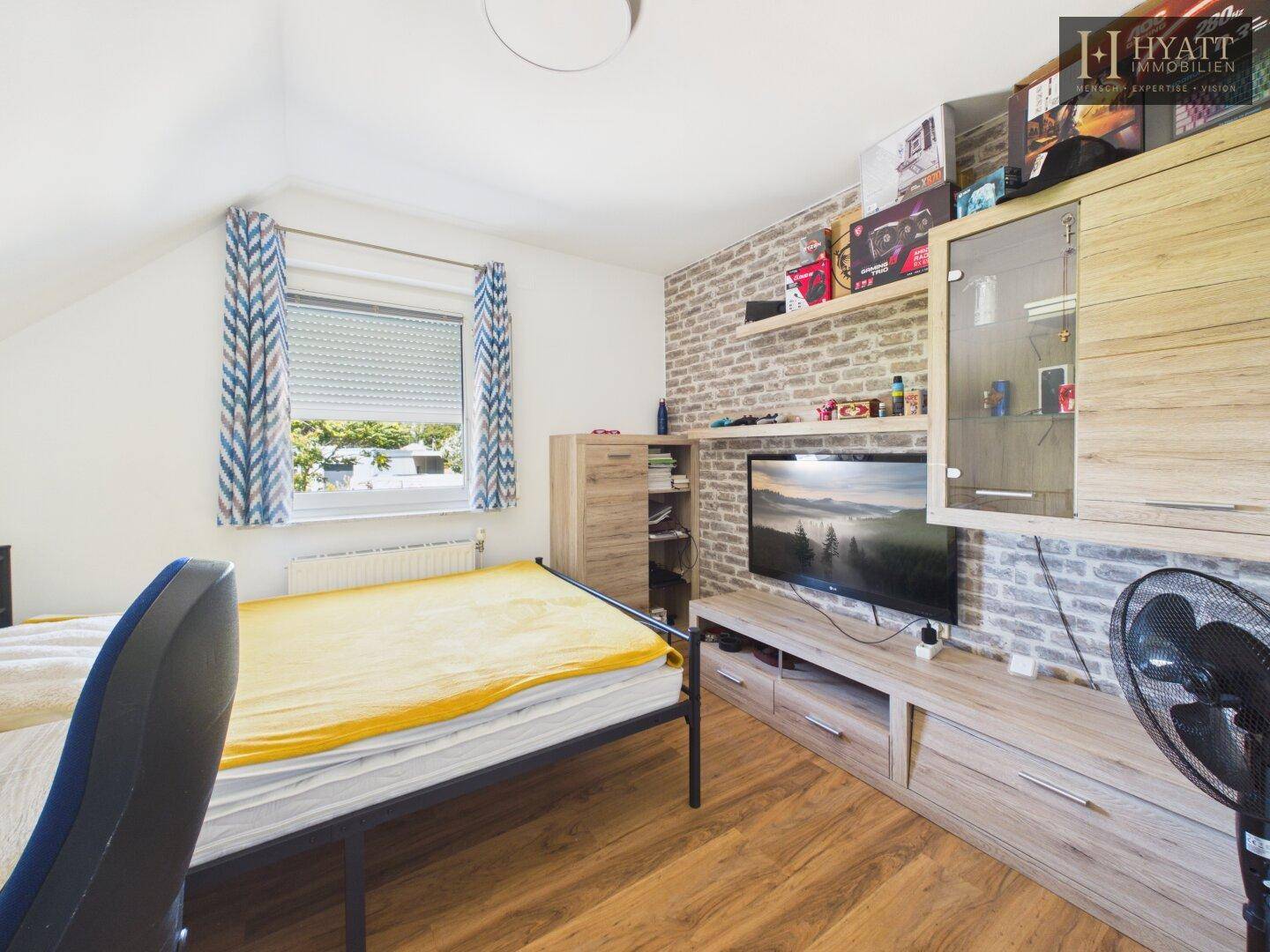 Schlafzimmer mit Einzelbett, Schreibtisch, Fernseher und Fenster mit Rollladen und Ziegelwand-Optik.