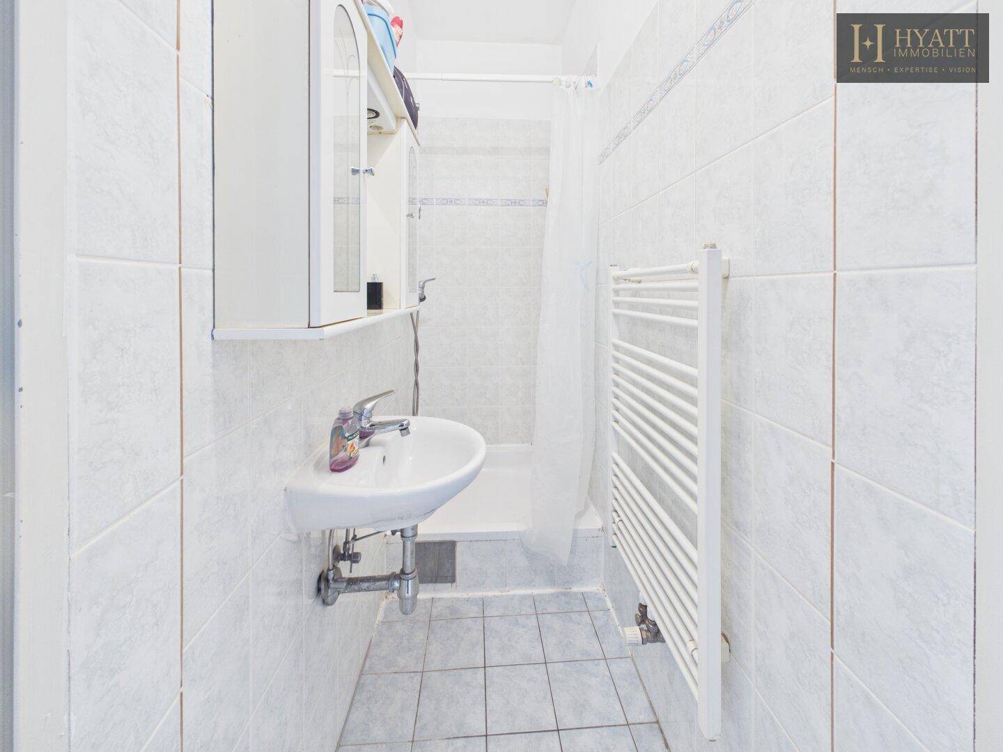 Helles Badezimmer mit weißen Fliesen, Waschbecken und Duschbereich.