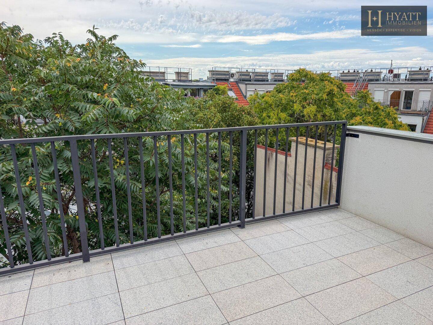 Leere Dachterrasse mit Fliesenboden und Geländer, umgeben von Bäumen und Stadtblick.