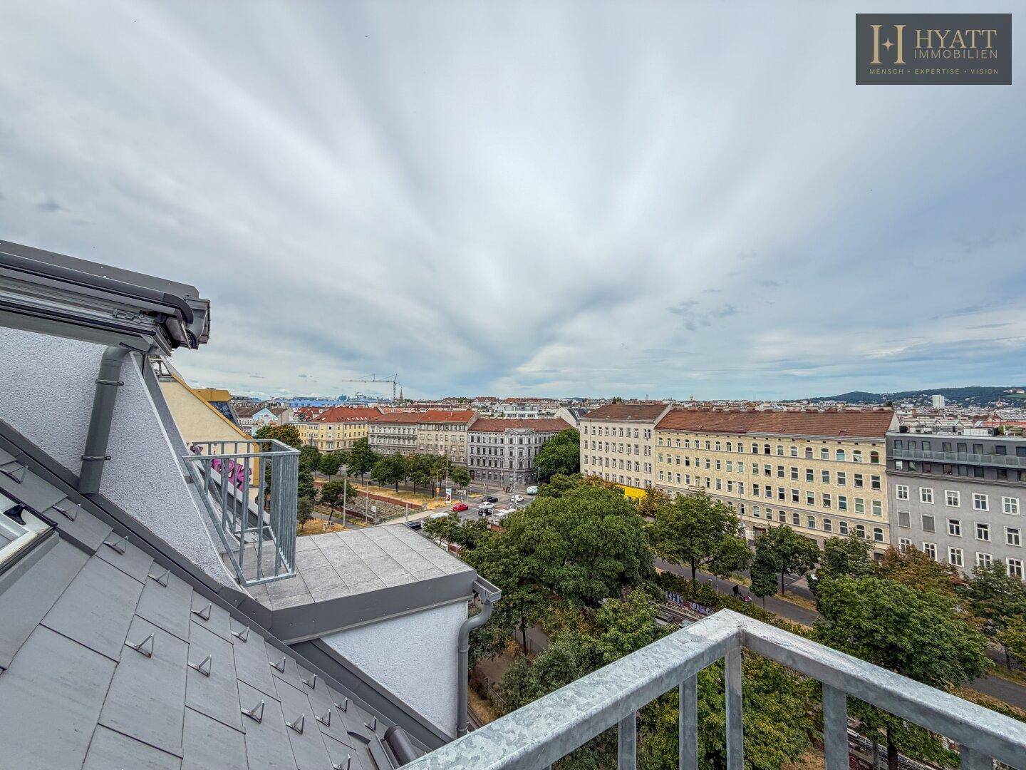 Balkon mit Blick auf die Stadt und die umliegenden Dächer, unter einem bewölkten Himmel.