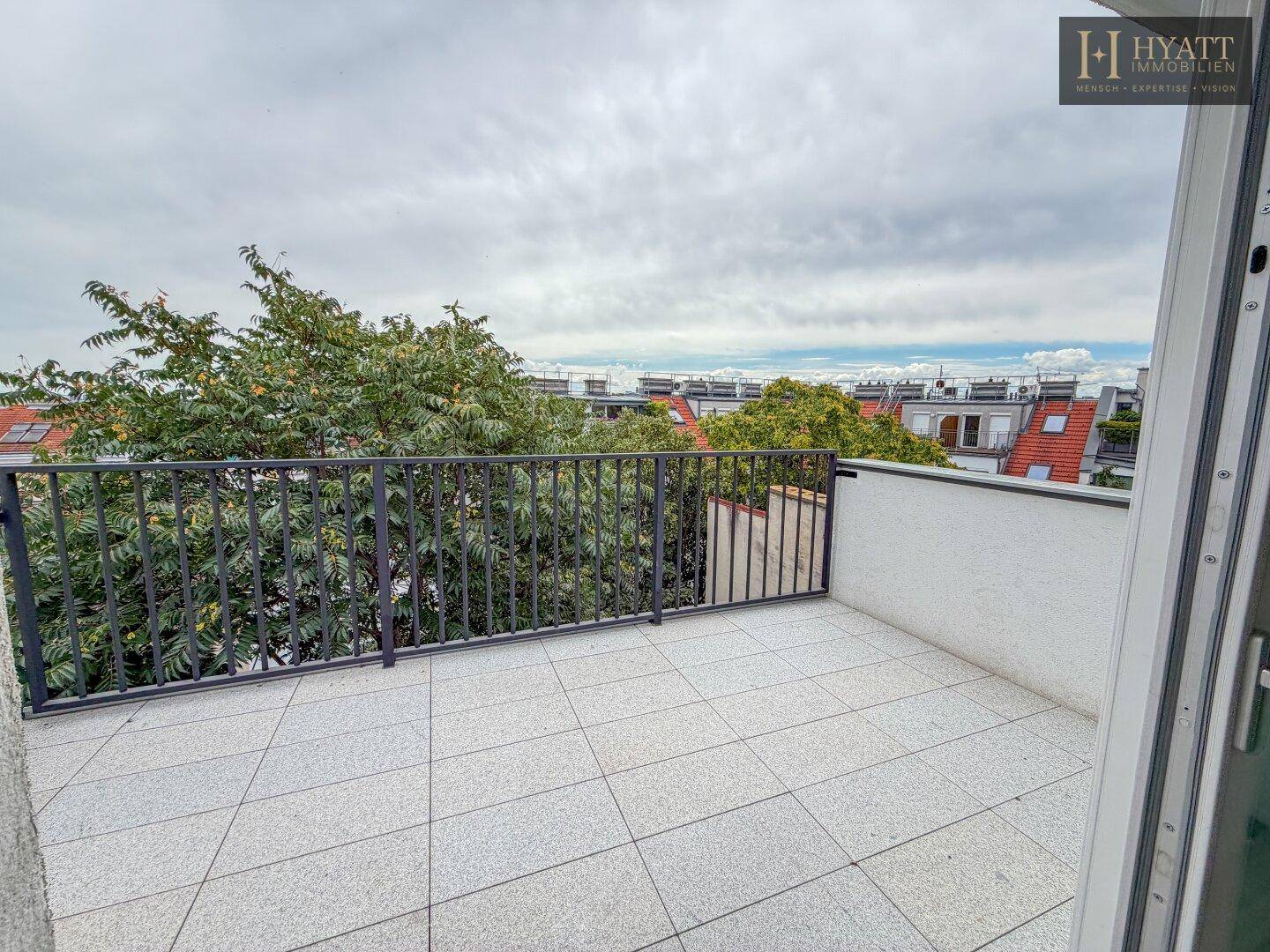 Geräumiger Balkon mit Fliesenboden und Metallgeländer, bietet Blick auf Bäume und Dächer.