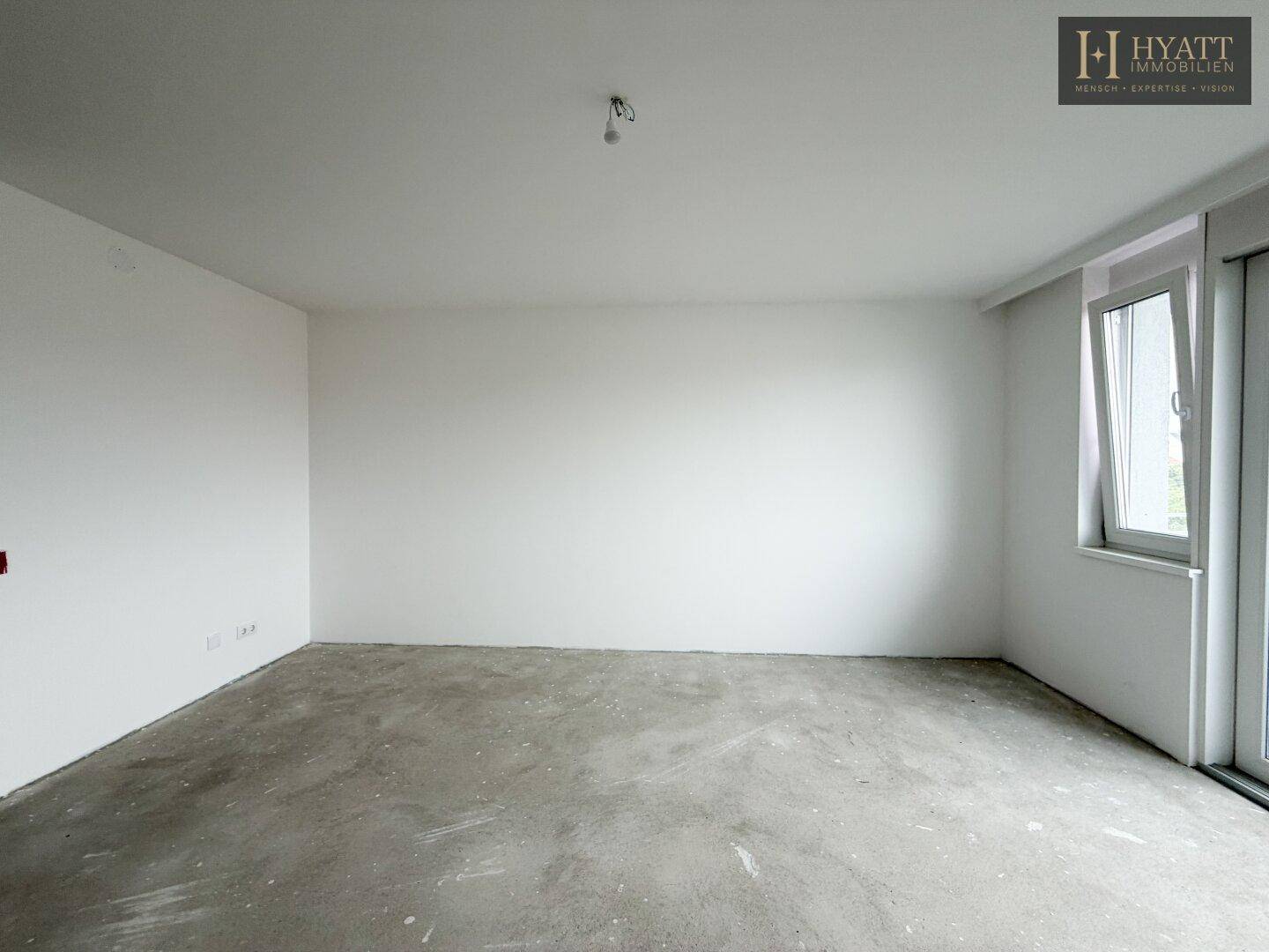Leeres Schlafzimmer mit Rohbetonboden und einem Fenster, das viel Tageslicht hereinlässt.