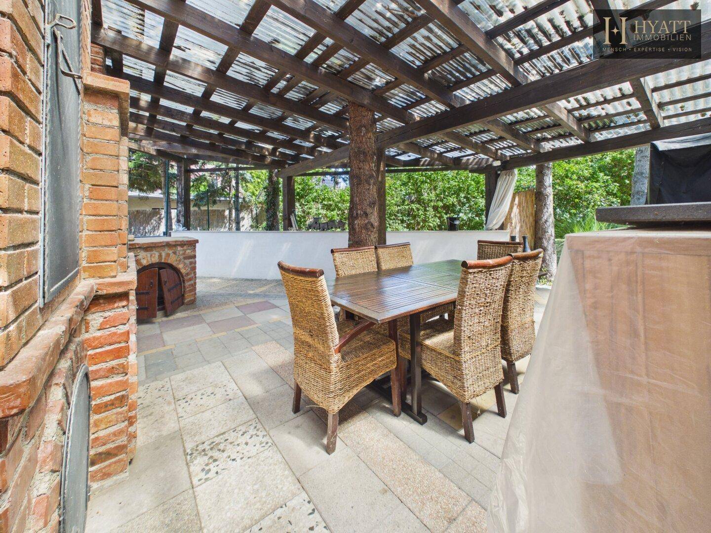 Geräumige überdachte Terrasse mit Esstisch, Korbstühlen und gemauertem Ofen für gesellige Abende.