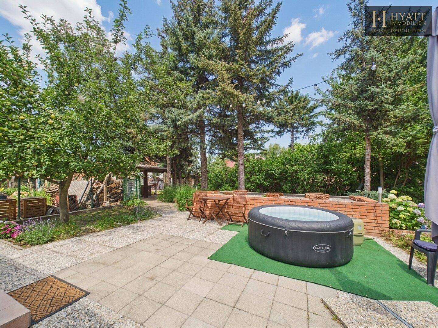 Gepflasterte Terrasse im Garten mit Whirlpool, Sitzgelegenheiten und umgebenden Bäumen.