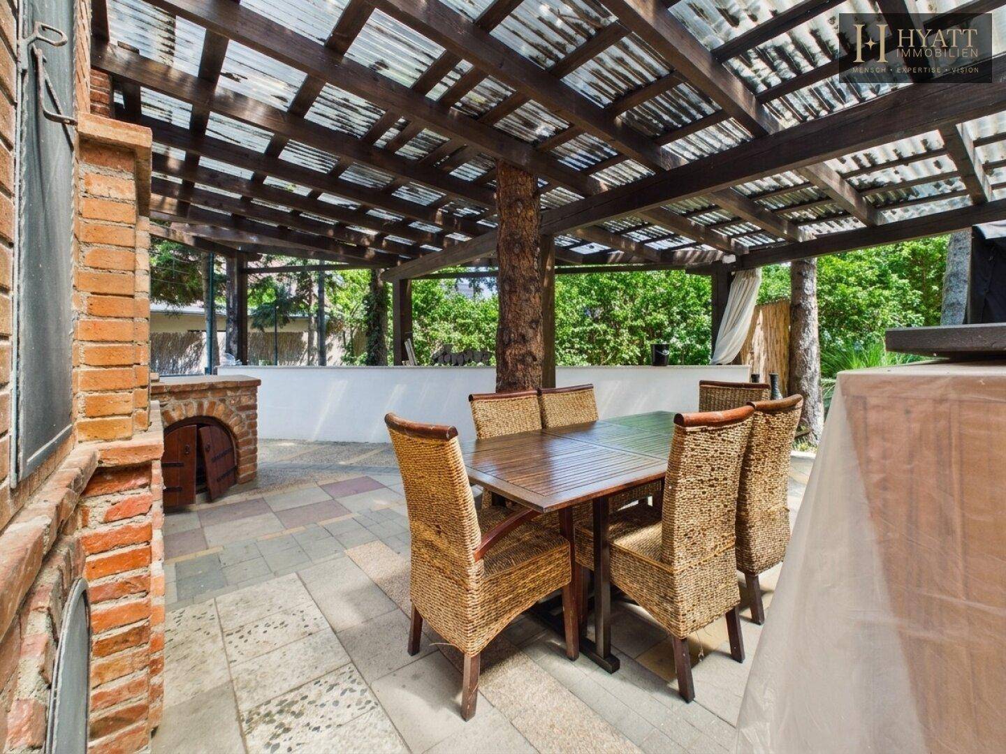 Überdachte Terrasse mit Essbereich und gemauertem Grill, umgeben von viel Grün.