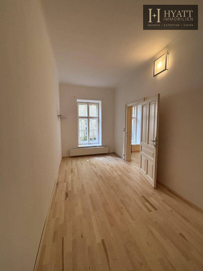 Leerer Raum mit hellem Holzfußboden, einem Fenster und einer geöffneten Holztür zu einem weiteren Zimmer.
