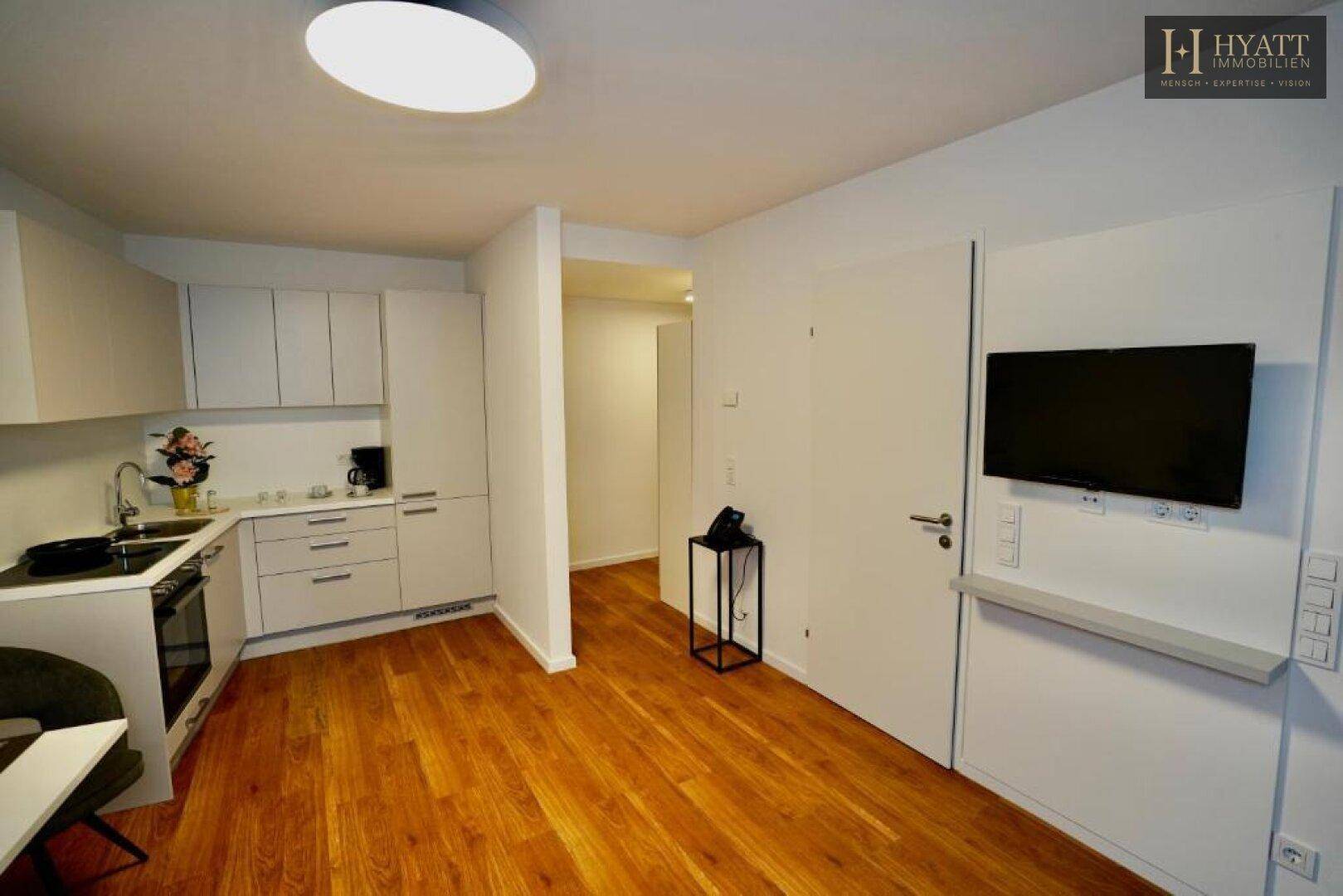 Moderne Küchenzeile mit weißen Schränken, Holzboden und Wandfernseher im offenen Wohnbereich.