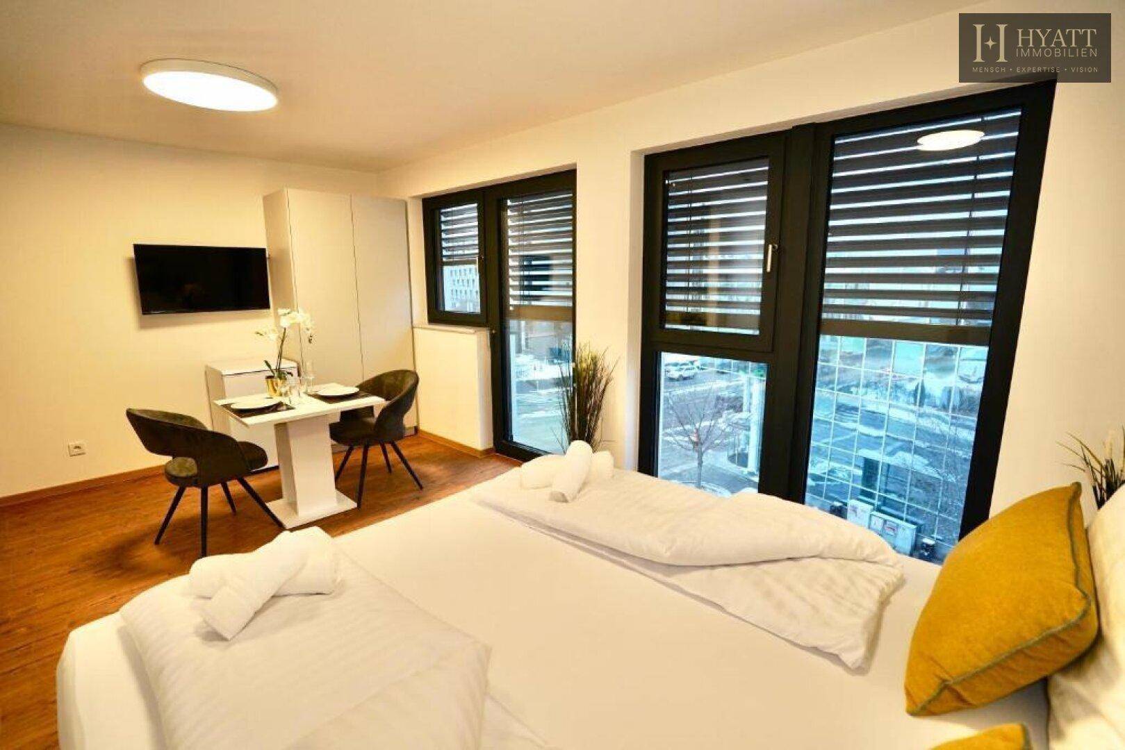 Schlafzimmer mit Essbereich, Wandfernseher und Blick auf die Stadt durch die Fenster.