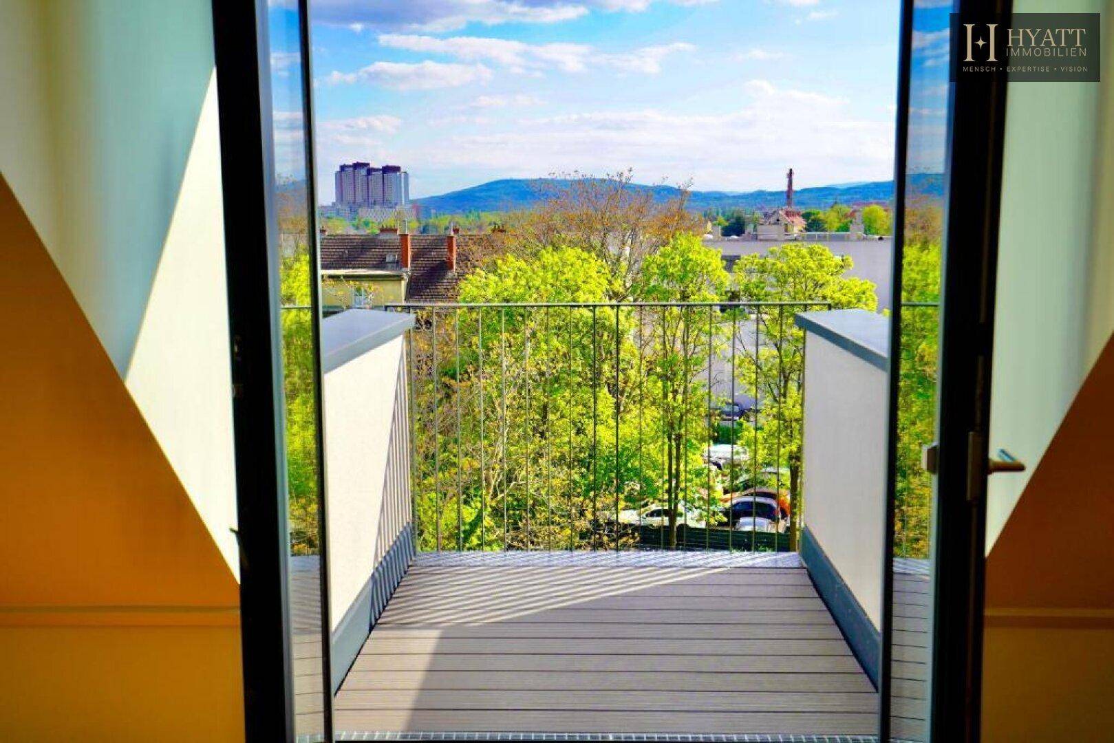 Kleiner Balkon mit Holzboden und weitem Blick über grüne Bäume und die Stadt.