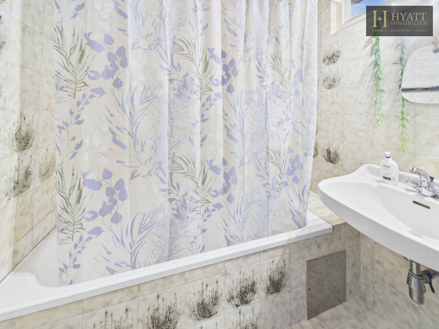 Helles Badezimmer mit Badewanne, Duschvorhang und älteren Fliesen.