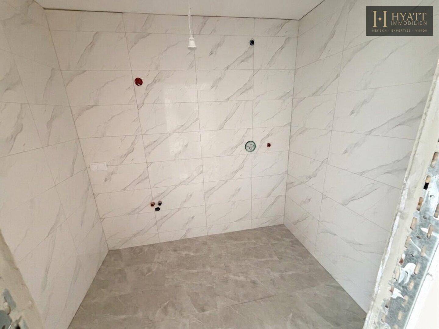 Badezimmer im Rohbau mit hellen Fliesen an den Wänden und vorbereiteten Anschlüssen für Armaturen.