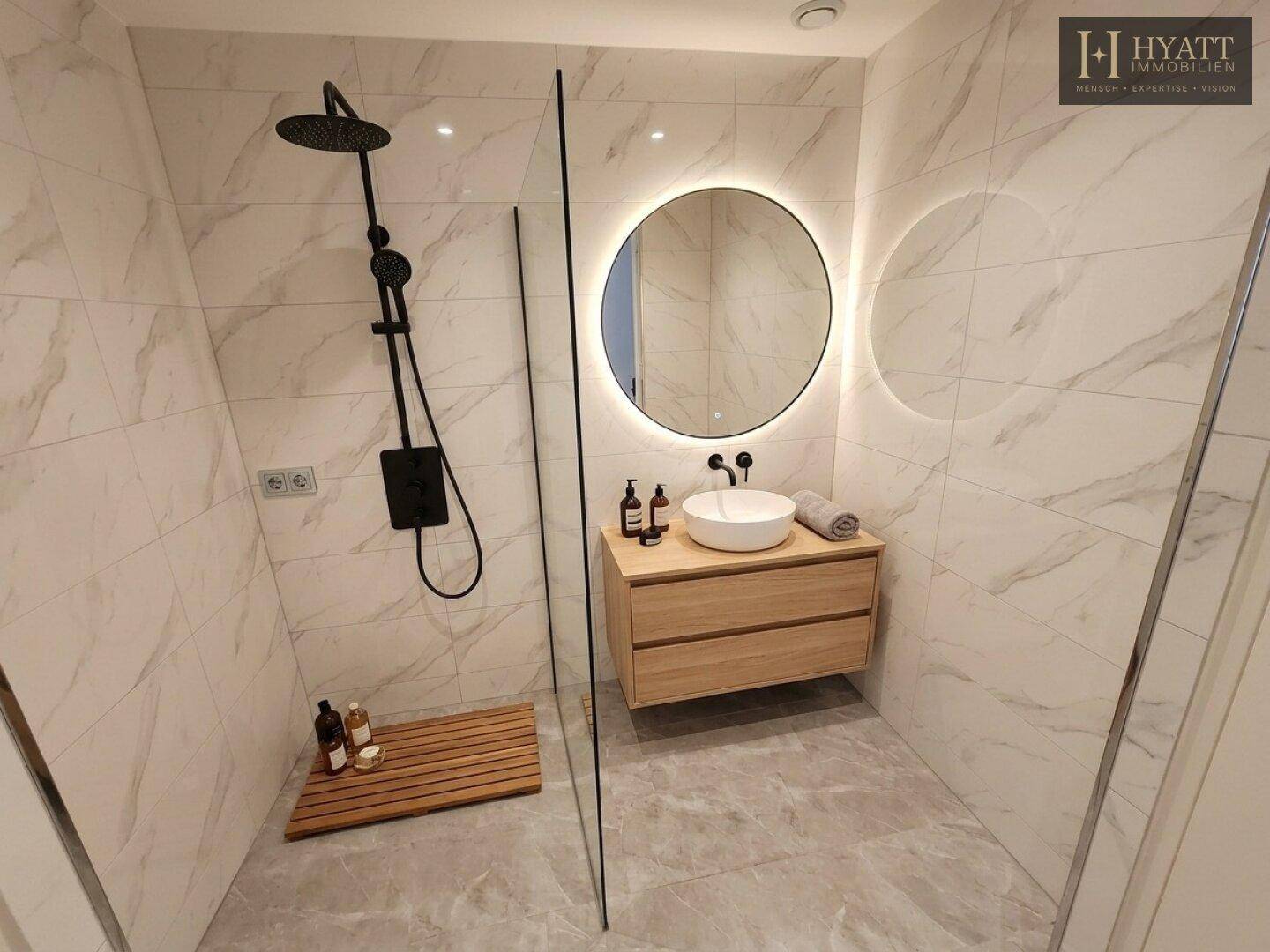 Modernes Badezimmer mit begehbarer Dusche, rundem Spiegel mit Beleuchtung und eleganten Fliesen.