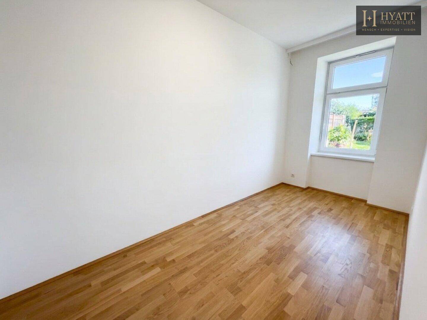 Leeres Schlafzimmer mit Parkettboden und großem Fenster, das einen Blick ins Grüne bietet.