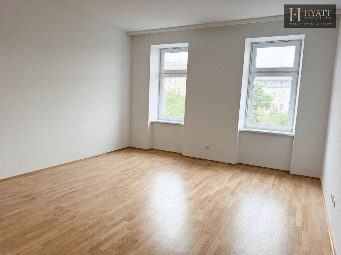 Helles, leeres Zimmer mit Parkettboden und zwei großen Fenstern, bereit zur individuellen Gestaltung.